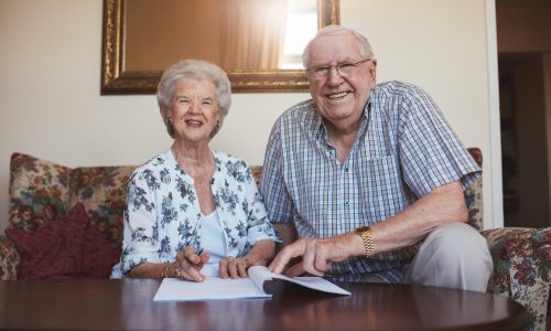 Portrait of a smiling retired couple looking over documents. Senior caucasian man and woman sitting on sofa at old age home and signing some paperwork.